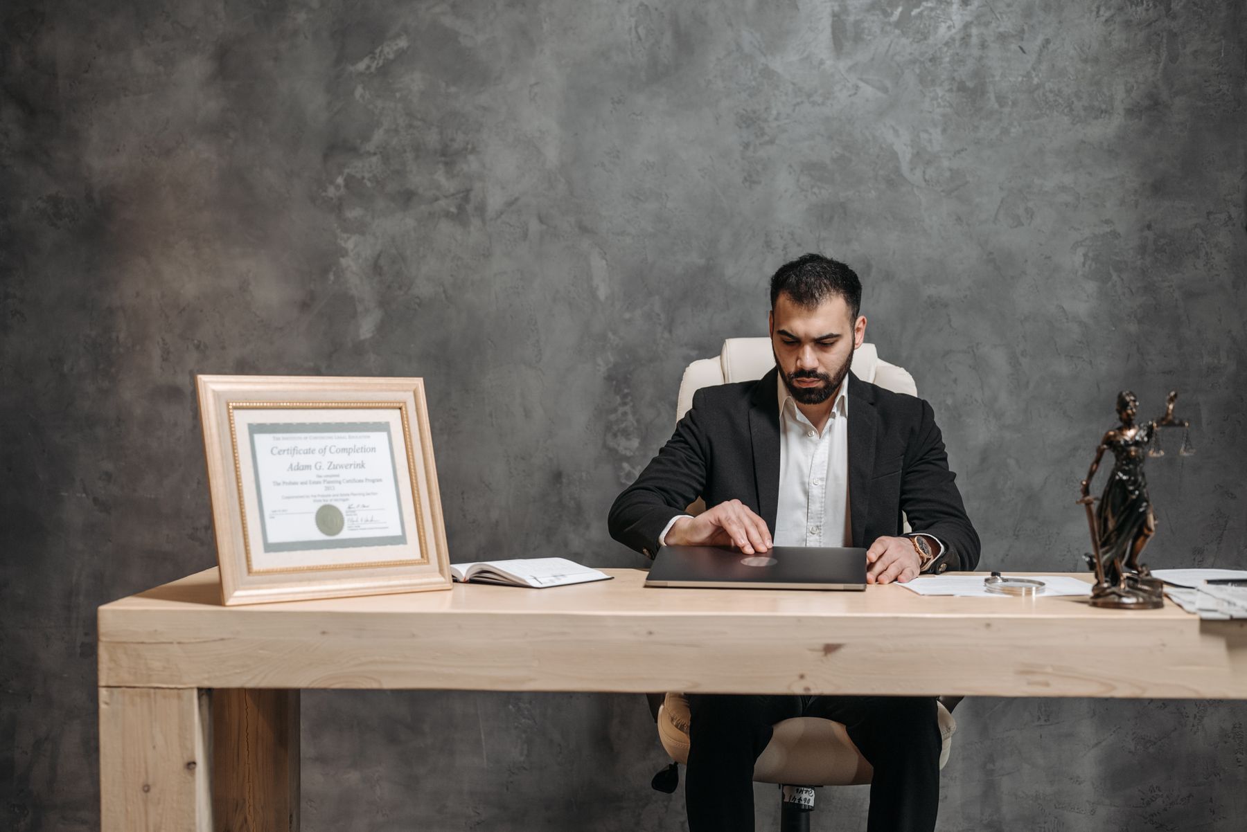 Advogado sentado à mesa do escritório, com balança da justiça, certificado emoldurado e processos ao lado do notebook